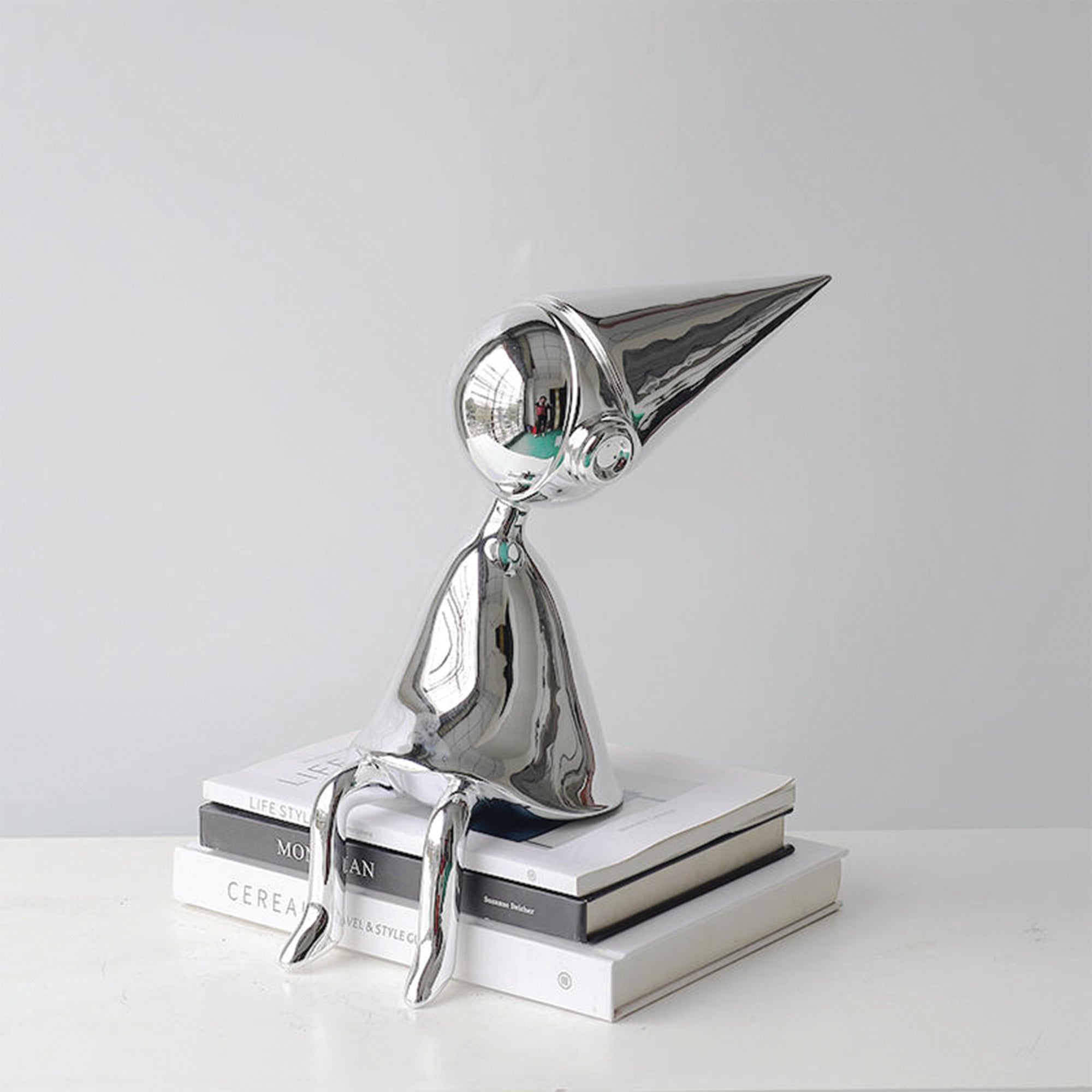 The Observer chrome sculpture by BAG&BONES displayed on stacked books, featuring a seated abstract figure with an elongated pointed hat and polished metallic finish.