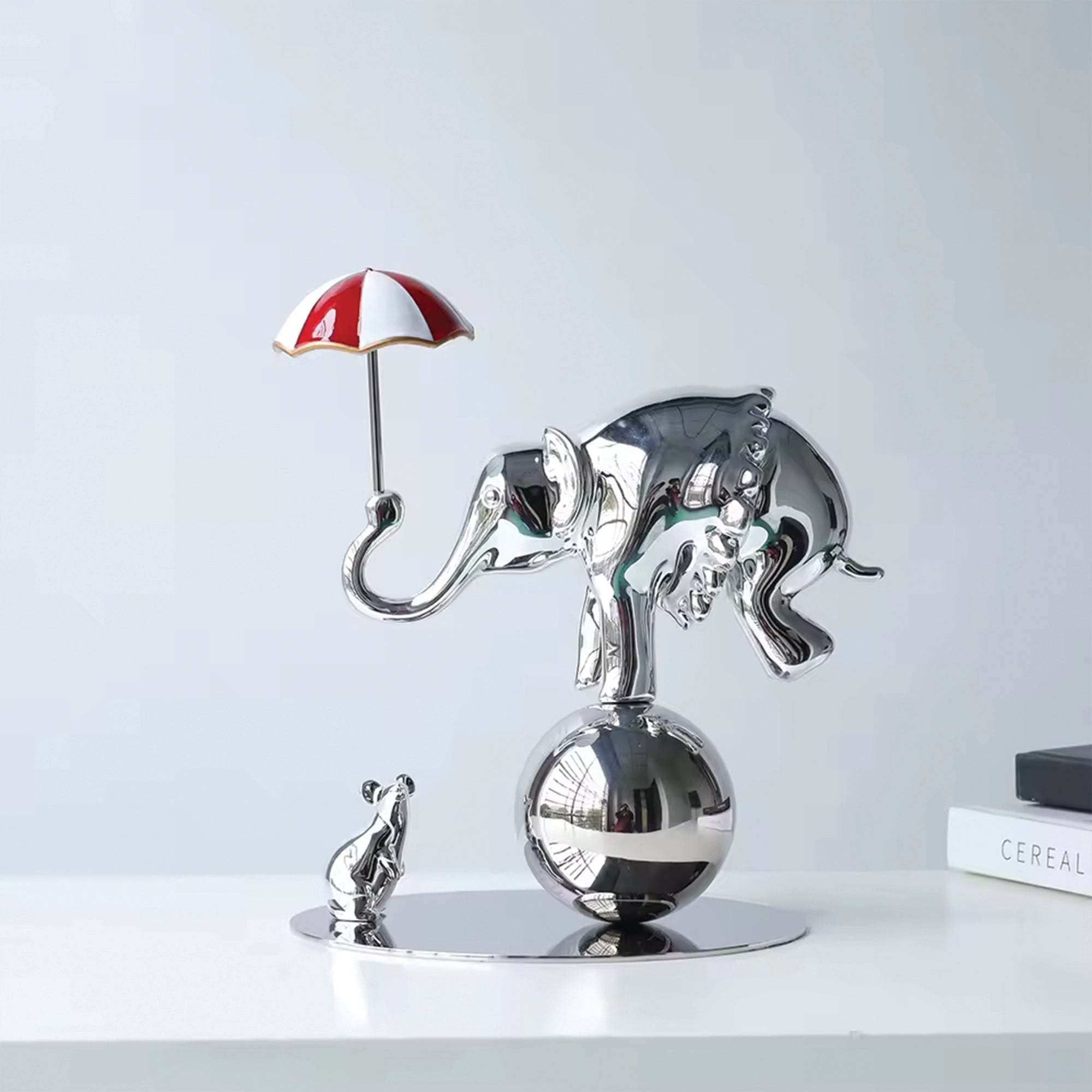 The Performer sculpture by BAG&BONES featuring a polished chrome elephant balancing on a sphere with a red and white umbrella and a small mouse below.
